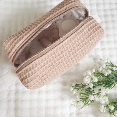 Estojo de tecido bege com padrão em relevo quadrado aberto sobre superfície branca acolchoada com flores ao lado