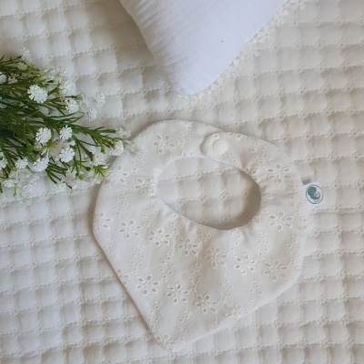 Babete branco bordado com botão sobre manta branca texturada com flores artificiais ao lado