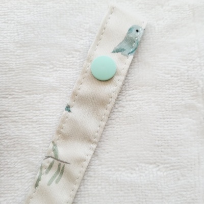 Faixa de tecido bege com botão azul e padrão de pássaros, sobre tecido branco felpudo