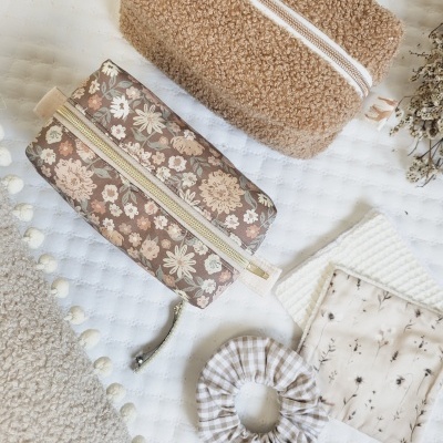 Bolsas de maquilhagem de tecido com padrão floral e felpudo castanho claro, elástico de cabelo às riscas e panos em tons neutros sobre uma superfície branca acolchoada.