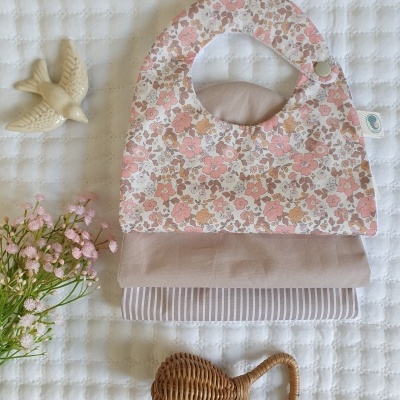 Três babetes de bebé empilhados em tecido floral, liso e riscado, com um chocalho de vime e flores cor-de-rosa num fundo branco acolchoado.