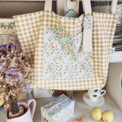 Saco tecido amarelo xadrez com bolso floral pendurado e necessaire floral em superfície com limões e flores secas.