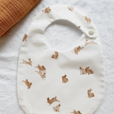 Babete branco com coelhos castanhos e fecho de pressão ao lado de manta castanha