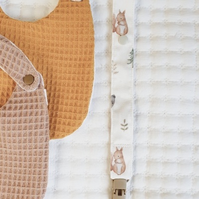 Dois babetes de tecido castanho claro e bege e uma fita para chupeta com desenhos de esquilos