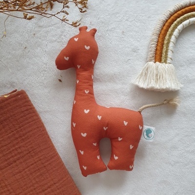 Brinquedo de pelúcia girafa com corações brancos sobre pano branco, junto a pano laranja e arco-íris decorativo de fios.