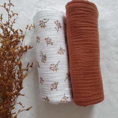 Dois tecidos enrolados, um branco com padrão floral castanho e amarelo e outro castanho, sobre superfície branca com ramo seco ao lado