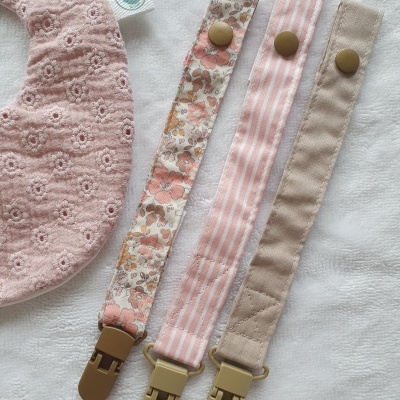 Três fitas para chupeta com padrões e babete rosa com padrão floral ao lado