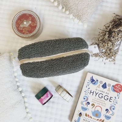 Bolsa cinza felpuda, livro HYGGE, recipiente vermelho, pequenos frascos, almofadas com pompons e ramos secos sobre toalha branca