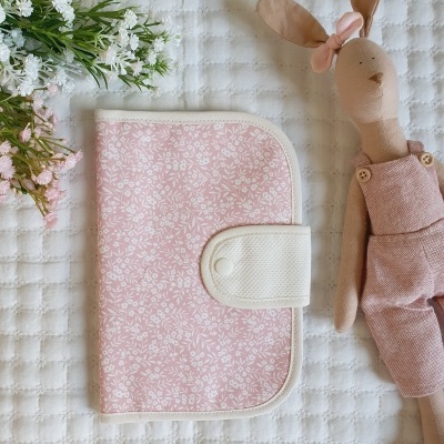 Capa rosa floral com fecho branco, boneca de pano rosa e flores brancas sobre tecido branco