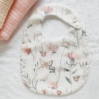 Babete branco com padrão floral e borboletas em tons pastel sobre toalha branca e toalhas dobradas ao lado