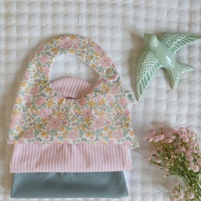 Tres babetes empilhados com padrões florais, riscado e azul, com um pássaro de cerâmica e flores ao lado sobre manta branca