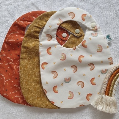 Três babetes de bebé com diferentes padrões e cores junto a um arco-íris decorativo de tecido