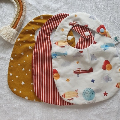 Conjunto de três babetes infantis com padrões distintos e cores variadas, disposto sobre tecido branco