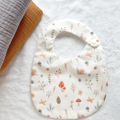 Babete branco com padrão de cogumelos, folhas e borboletas, fecho botão de pressão, sobre toalha branca e mantas cinzenta e castanha.