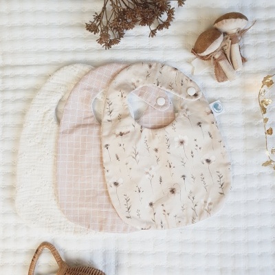 Três babetes com padrões diferentes sobre fundo branco com flores secas e brinquedo de vime