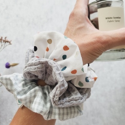 Mão segurando frasco de spray para tecidos com três scrunchies coloridos no pulso