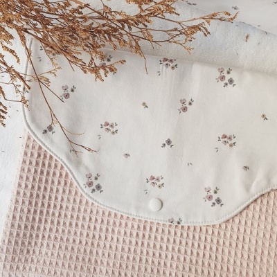 Babete branco com pequenas flores sobre pano rosa texturizado e ramos secos.