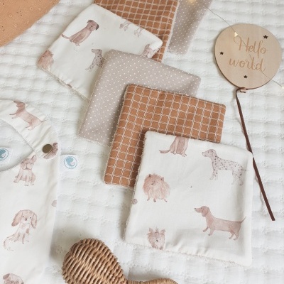 Panos de algodão com motivos de cães em tons neutros, brinquedo de vime e balão de madeira com texto Hello world sobre superfície branca acolchoada