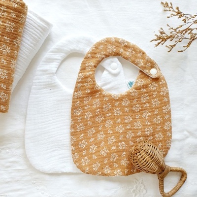 Três babetes de bebé com padrão floral e textura, brinquedo de madeira e ramo seco sobre fundo branco.