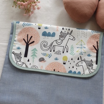 Bolsa ou manta azul com aba estampada de unicórnios e árvores sobre superfície branca com flores e almofadas tons terra