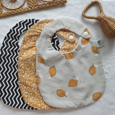 Três babetes para bebé com padrões em preto e branco, amarelo e branco e cinza com limões, junto de chocalho de palha