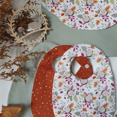 Três babetes estampados com padrão de esquilos e folhas e desenho de bolinhas brancas, sobre tecido verde claro e branco com decoração em madeira e ramo seco