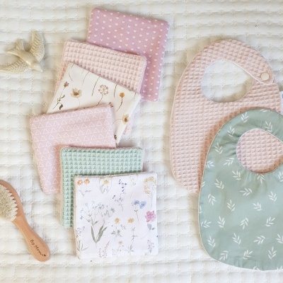 Conjunto de produtos para bebé com babetes, panos, escova de cabelo e acessórios em tons pastel e padrões florais sobre superfície branca