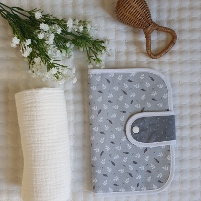 Estojo dobrável cinzento com padrão de folhas, toalha branca enrolada e espelho de mão em vime castanho sobre tecido branco