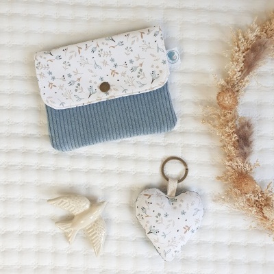 Bolsa azul com aba floral, chaveiro coração floral e pássaro de cerâmica sobre superfície clara texturizada, com flores secas ao lado.