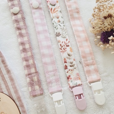 Cinco fitas coloridas com padrão de quadrados e floral, cada uma com botão e clip plástico, sobre pano branco com flores secas.