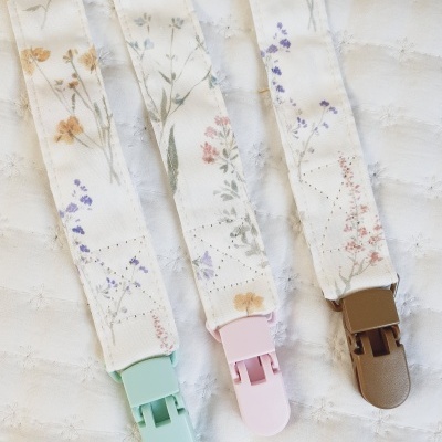 Três prendedores de chupeta florais com clip de plástico colorido sobre tecido branco acolchoado