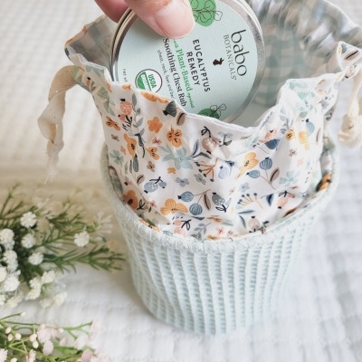 Recipiente metálico com pomada dentro de saco de tecido floral e base azul, sobre superfície branca com flores