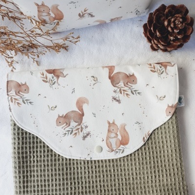 Saco de tecido com padrão de esquilos e folhas secas, fundo branco e parte inferior verde oliva texturizada, com pinha e ramo seco ao lado.