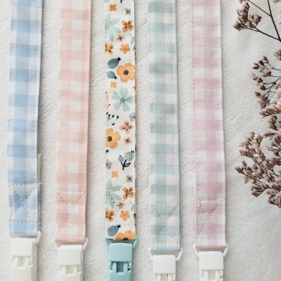 Cinco fitas para chupeta com padrões às quadrículas e uma com padrão de flores, com clips de plástico.