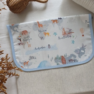 Produto têxtil com mapa-múndi ilustrado, bordas azuis, sobre tecido bege e decoração com vaso de junco e flores secas