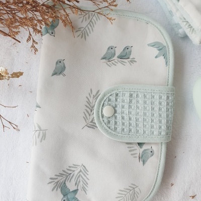 Capa branca com padrão de pássaros azuis e ramos verde-claros e fecho de botão de pressão com textura quadriculada.