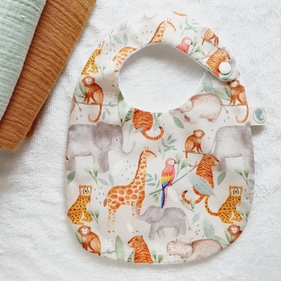 Babete com padrão de animais da selva em tecido, sobre toalha branca, perto de duas toalhas dobradas castanha e verde clara.