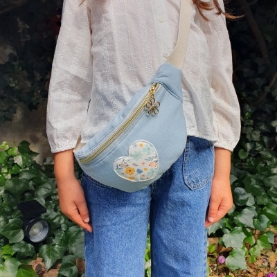 Mini bag azul clara com padrão de coração florido e fecho dourado, alça bege, usada por pessoa com camisa branca e calças de ganga azuis.