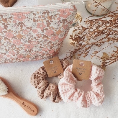 Dois scrunchies de tecido castanho e rosa claro com etiqueta, escova de madeira 'By Mom' e bolsa floral