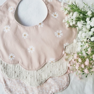 Três babetes de tecido com padrões de flores colocados sobre tecido branco com flores frescas ao lado