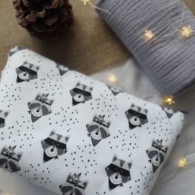 Tecido branco com padrão de guaxinins cinzentos e pretos, rolo de tecido cinza claro, pinha castanha e luzinha decorativa