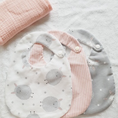 Três babetes de tecido com padrões de ovelhas e listras em diferentes cores e um pano rosa claro num fundo branco texturado.