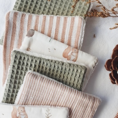 Toalhetes infantis em algodão com padrões verdes, beges e estampas de esquilos, dispostos sobre tecido branco com elementos naturais ao lado.