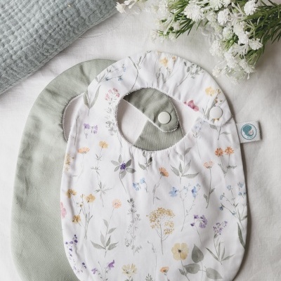 Dois babetes de bebé, um verde e outro branco com flores, junto a flores brancas e almofada azul