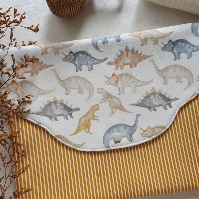 Manta ou fralda com padrão de dinossauros e riscas amarelas, em superfície clara com brinquedo de madeira e toalha branca