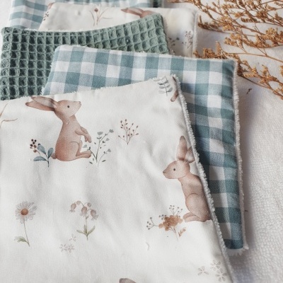 Conjunto de três panos de tecido com estampas de coelhos, padrão xadrez azul e branco, e textura verde sobre superfície branca e ramo seco ao lado.