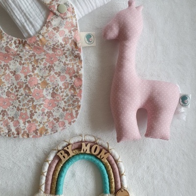 Babete floral, peluche de girafa rosa e enfeite arco-íris com letras BY MOM sobre tecido branco