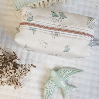 Necessaire branca com padrão de pássaros e pássaro decorativo verde em cerâmica sobre manta branca.