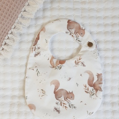 Babete branco com padrão de esquilos castanhos e folhas sobre manta branca texturizada e tecido castanho com franjas