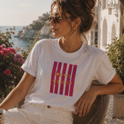 Mulher com t-shirt branca com riscas rosas e texto amarelo sentado em cadeira ao ar livre com mar e casas ao fundo